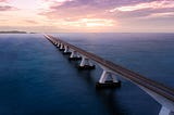 A two-lane highway stretching away over a body of water, toward a sunset in the distance.