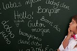 A young girl looking pensively at a chalkboard with various foreign translations of the word ‘hello’ written on it