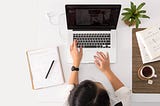 A person using a laptop surrounded by a notepad, plant and coffee