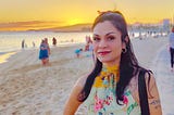 This image shows a person at a beach during sunset. They’re wearing a colorful floral-patterned top with yellow, pink, and green elements. The background features a beautiful golden-orange sunset over water, with other beachgoers visible in the distance. The beach has light-colored sand, and the atmosphere appears warm and peaceful. The person is smiling at the camera and has dark hair, hoop earrings, and appears to be enjoying the scenic beach environment.