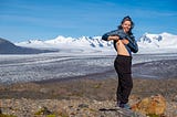 Amanda is standing in front of a Patagonian mountain range and lifting up her shirt and jacket to reveal a sunflower tattooed on her ribs.