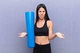 Young woman holding yoga mat , arms out, funny look on her face