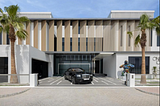 A cubist-inspired modern mansion with white walls, large square windows and large louvred panelling on the top floor. In the driveway sits a black Rolls Royce. To the right of the Rolls, there’s a graffitied statue of a gorilla standing on a plinth. Blue skies and tall palm trees indicate that this house is somewhere hot.