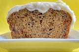 Image of banana bread on a white plate. a piece has been cut off from the end revealing the inside. the banana bread is frosted with a cream cheese frosting. it is sitting on a white plate. the background is bright yellow.