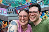 A photo of myself (a smiling white woman with pink glasses, brown hair, and tattoos) and my husband (a smiling white man with brown hair and glasses wearing a green shirt. We are sitting in front of a colorful mural of houses and trees