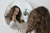 A brunette, Caucasian woman ponders in front of a mirror.