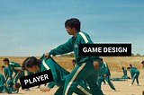 Homem de pele morena com cabelo escuro vestindo uniforme de moletom verde com listras laterais brancas, com a tag “game design” segurando outro homem de pele amarela e cabelo escuro, vestindo o mesmo uniforme, com a tag “player” em meio a um campo de areia com outras pessoas ao fundo com o mesmo uniforme.