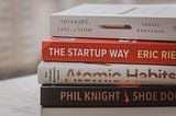 Close-up on a stack of four self-help books, laying on their sides on a table. Titles include “Thinking Fast and Slow,” “The Startup Way,” “Atomic Habits,” and “Shoe Dog.”