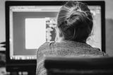 Woman with blonde hair in a bun facing away toward a computer screen.