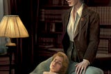 A digital painting of Eleanor wearing a tailored tweed brown suit, her brunette hair in a bun, and round gold-rimmed spectacles, standing over the sea-green chaise lounge that Eddie is lying on. Eddie has short blond hair and is wearing a white shirt. The background is a dimly-lit office with bookshelves and one floor lamp.