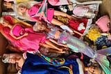 The image features a cardboard box full of Barbie dolls and a few Ken dolls. In the foreground, there’s a mermaid Barbie with a purple fishtail, a rockstar Barbie holding her pink guitar, a Hawaiian Barbie not in her original clothes, a princess Barbie and a Mulan Barbie.