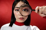 Picture of a woman with shoulder length dark hair standing in front of a red background. She is looking at you through a magnifying glass.