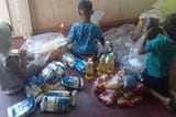 Three children sit on a floor with their backs to the camera surrounded by food goods from a humanitarian food basket.