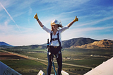 A blonde woman stands on top of a wind turbine with her arms raised