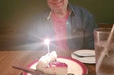 man sitting with cake + candle at restaurant table