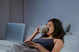 woman in blue spaghetti strap top reclining on bed using laptop and drinking