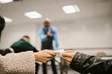 Two students passing notes in a classroom who are caught by a teacher.