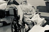 This is a sepia-toned photo of a nursing school manikin, that was taken in a medical learning facility/lab. The manikin has long shaggy hair, a hospital gown, and is sprawled out in a wheelchair in a very uncomfortable and unnatural looking position. The mood seems ominous.