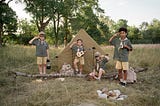 Group of children dressed as scouts at a campsite. They are surrounded by camping gear with a tent set up behind them. One child is playing the ukulele, another is reading a book, while the others are saluting. They are outdoors in a grassy area with trees in the background.
