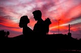 Photo of a man and woman’s silhouettes looking at each other, with a purple and red sunset sky behind them.