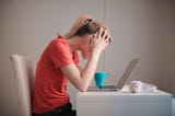 A woman wearing an orange t-shirt sits over a laptop computer, holding her head in her hands as if overwhelmed.