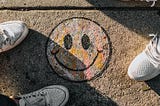 Smilie face drawn with chalk on the foot path with people’s feet in sneakers around it.