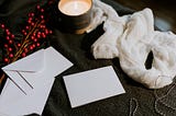 Photo on a dark background of a table, with white envelopes and note cards in the center of the photo. A white scarf lays above the cards, to the right of the photo. A candle in a tin is lit, to provide ambience. A bough of red berries decorates the table.