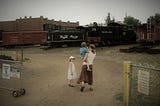 A young woman with a small boy on her hip and a young girl to her left, walking into an old train yard. The woman and boy have their backs facing, while the girl is turned to the right side. A black train engine in the background reads “Royal Gorge”.