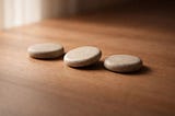 Three smooth, flat stones arranged in a horizontal line on a warm wooden surface, with the center stone slightly rotated, softly lit with shallow depth of field and blurred background.