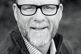 Black and white picture of an older man who is bald and wearing glasses and has a beard with gray hair.