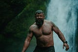 Candid photo of a tattooed bearded man in water next to a waterfall