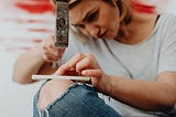 A woman about to try hitting a wood screw with a hammer into a thin piece of wood balanced on top of her leg