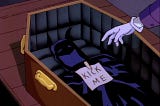 A screenshot from ‘Batman: The Animated Series’. The Joker’s hand can be seen attaching a “kick me” sign to Batman’s cape and cowl, inside a coffin.