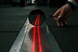 A red line of lights running towards a mirror, which reflects them at a slightly different angle. A hand holds the mirror. The background is a dark street.