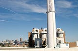 A large white smokestack with Baltimore written on it is silhouetted against a blue sky.