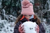Little girl bundled up in a knit hat and gloves holding a snowman’s head-sized ball of snow. She looks cold!