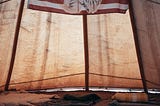 An image of a Native American elder imposed in the middle of an United States flag hangs from the inside of a tent styled in the tradition of an Indigenous tribe, complete with fur pelts, blankets, and a stone bordered fire pit on the earth beneath.