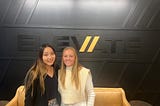 Davidson College students Marie Ueda ’25 and Ava Blasch ’26, dressed in business casual attire and smiling at the camera, stand in front of a beige couch, small brown table, and black wall with the word “ELEVATE” written on it.