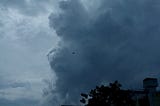 Dark, dramatic storm clouds loom over a cityscape in the early evening, casting deep shadows over buildings and trees.