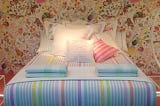 A picture of the Zara Home shop, with a bed with white bedlinen, with colorful stripes (purple, blue, grey, yellow, green, pink and orange). Lots of white cushions complete the bed.