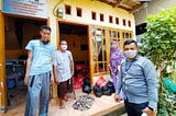 4 people stand in front of a house-like structure. Two people stand on either side of the photo, and bags of food deliveries