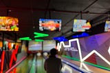 A person, how hands are hidden in front of them, stands just before an open lane at a bowling alley. All pins are set for a fresh take at the bowl. Neon lights are all around, the room feels dark and cool. It is ready to be played.