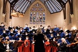 A medium-sized choir of adults and children stands in formation, in choir robes, poised to sing with a chamber orchestra in a church; you see the back of the conductor, poised to conduct.