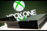 A stylish promotional image of a sleek rectangular video game console and its motion sensing camera accessory. The words “Xbox One” are blurred in the background.