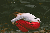 A person wearing a white long-sleeve top and red pants crouches on the ground in a forest, head down and arms wrapped around their knees, surrounded by fallen leaves and dark green foliage, conveying a mood of sadness or introspection.