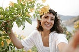 A photo of a woman with dark hair and light brown skin smiling next to a tree with yellow flowers.