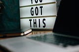 “You Got This” light sign next to a journal and laptop that are blurry