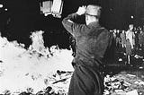 A black and white, grainy photo of a pile of books in the street, on fire. A crowd is visible looking on, and in the foreground, a Nazi soldier facing away from the camera throws several more books onto the pile.