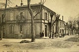 Black and white picture of the post office in Medea, this is a two story European style building on a tree-lined boulevard. A group of people, including men in suits and girls in white dresses is seen posing in front of the building.