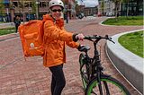 A woman standing next to a bike wearing a helmet, an orange jacket and a large orange backpack meant to transport food orders.